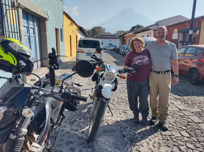 Two happy people stand on a cobblestone street. Two motorcycles are in front of them. There is a volcano far in the background. 