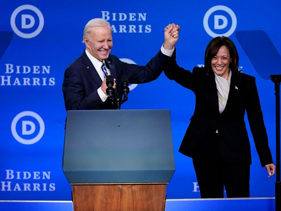 Biden and Harris are holding hands, in a triumphant manner, behind a podium and in front of a backdrop that says Biden Harris.