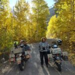 A woman stands between two motorcycles, amid trees that are bright yellow