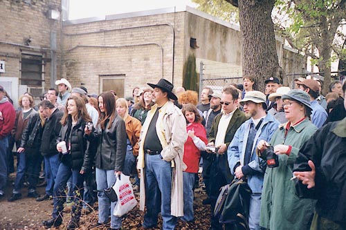 A large group of people are standing and looking at something out of the photos frame, except for one woman in the middle, who is looking at the camera, has a dorky expression on her face and is giving a thumbs up. 