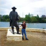 A woman stands next up to Stevie Ray Vaughan statue on the Austin, Texas water front. She is smiling. There are rowers behind her on the river.