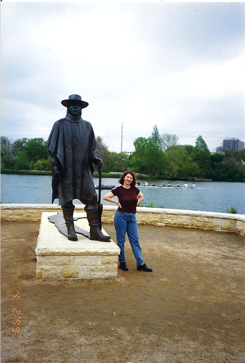 A woman stands next up to Stevie Ray Vaughan statue on the Austin, Texas water front. She is smiling. There are rowers behind her on the river.