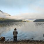 a woman stands with her back to the camera. She is looking out over a beautiful lake. She is alone.