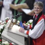 A smiling Bishop stands at the church lectern, which is covered in flowers.