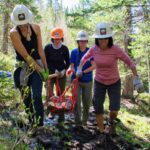 Four women on a trail in the woods carry a large rock in a net.