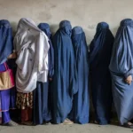 A line of women, all but one in a blue burqa, stand in line.