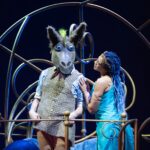 a man has a large fake donkey head on his head. A beautiful woman is touching him, and looks like she is in love with him.