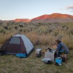 A tent is set up in a remote, high desert landscape, with a woman nearby sitting on a motorcycle pannier, at a table assembled from panniers.