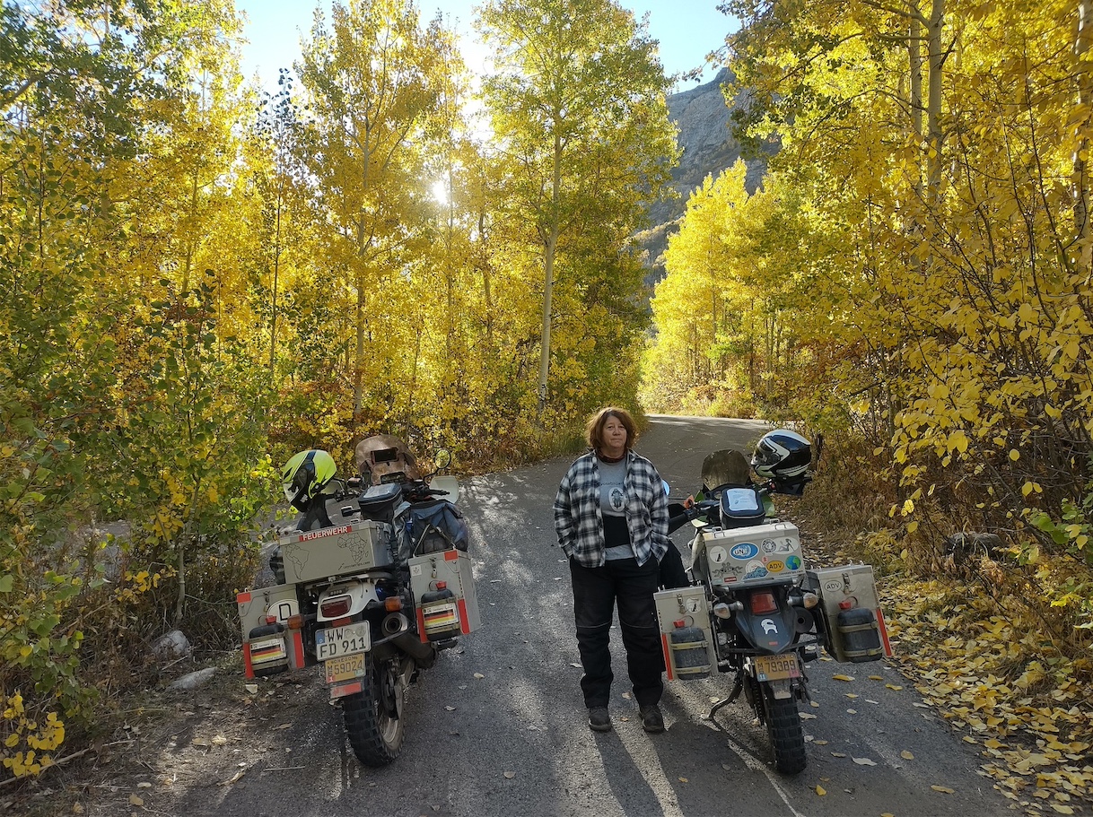 woman stands between two motorcycles in a scene filled with
        golden leaves