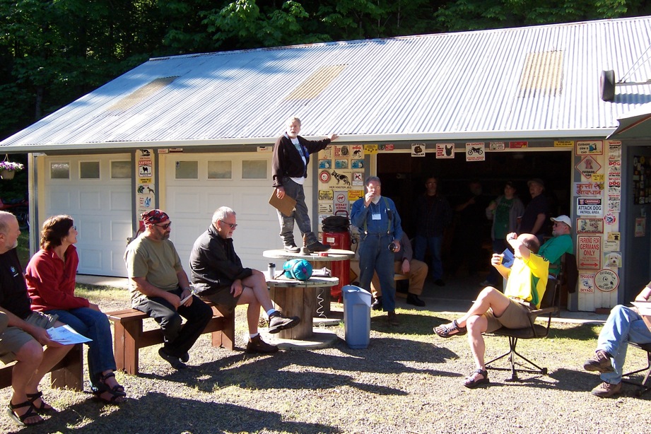 A man stands
        on a table and addresses people sitting around him. They are
        outside.