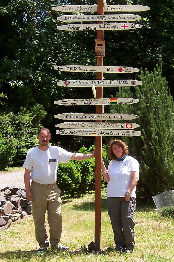 Two people stand
        in front of a pole that has slats on it, and each slat has the
        name of a traveler or travelers that visited Eric and Gail's in
        Westfir, Oregon.